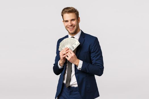 Business, finance and money concept. Cheerful, pleased handsome blond bearded businessman in classic suit, tie, holding lots cash, winning bid, smiling pleased, standing white background.
