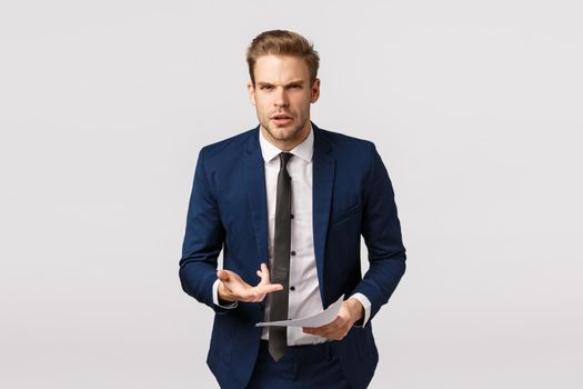 Furious, aggressive young businessman in classic suit, frowning staring outraged and pointing at documents pissed, scolding employee writing bad report, standing bothered white background.