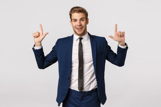 Surprised and glad, excited smiling bearded businessman, male entrepreneur in suit, showing product banner, pointing fingers up and look camera with delighted grin, standing white background.