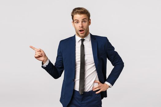 Surprised, speechless young businessman with blond hair, bristle, wearing classic suit, starting career new company, asking question about coworker, pointing left and look amused, white background.