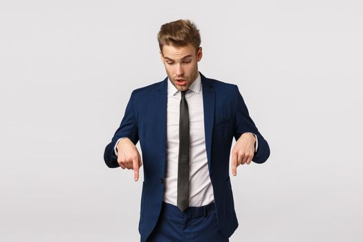 Shocked, impressed and curious blond bearded guy in classic suit, businessman pointing looking down speechless, standing astonished and impressed white background, check out product banner.