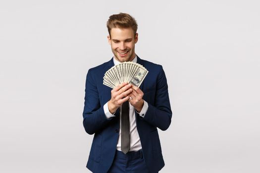 Happy and excited, good-looking businessman in classic suit, counting money, holding big cash dollars and smiling with pleased, delighted expression, standing white background glad.