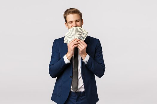 Finance, economy and business concept. Attractive successful young businessman in suit, holding lots of cash, sniff dollard celebrating achievement, signed good corporate deal, white background.