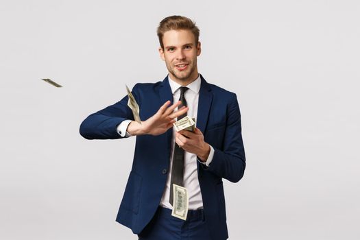 Success, money and finance concept. Handsome confident, blond bearded businessman in suit, holding cash and throwing money in air with pleased, satisfied expression, wasting dollars.