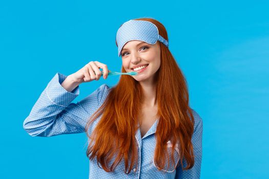 Cheerful cute redhead woman in nightwear, pyjama and sleep mask, brushing teeth holding toothbrush and smiling cheerful, wake up and taking care hygiene, standing blue background.