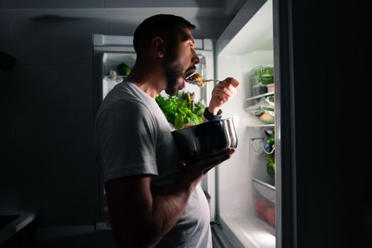 Young hungry man eating food at night and looking in open fridge. Man taking midnight snack from refrigerator. High quality photo
