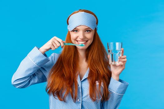 Enthusiastic upbeat, lively redhead woman, foxy girl with pretty smile, brushing teeth holding toothbrush and glass water, wake up morning, getting dressed, wearing sleep mask and pyjama.