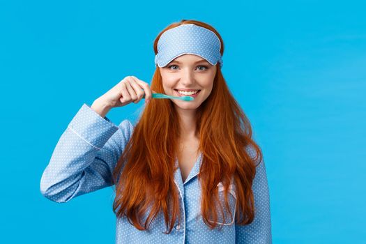 Habits, hygiene and lifestyle concept. Cute feminine redhead european woman in nightwear, sleep mask smiling and brushing teeth with toothbrush, standing blue background.