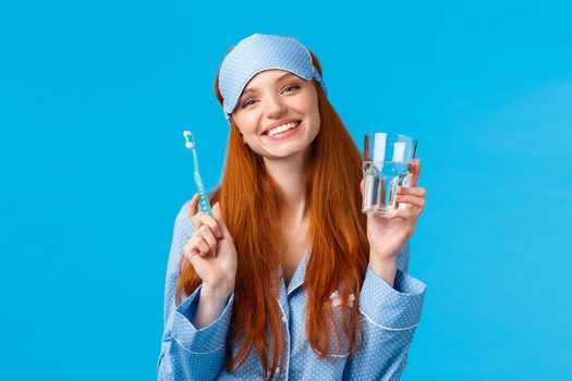 Happy smiling and carefree redhead enthusiastic girl in sleep mask, pyjama, holding glass water and toothbrush, taking care mouth hygiene, dental daily routine, blue background.