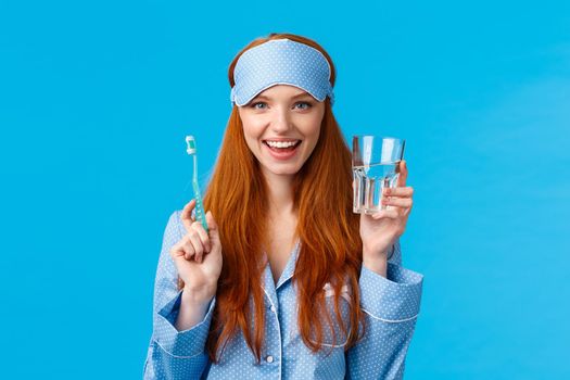 Advertising, hygiene and people concept. Feminine cute redhead girl feeling upbeat and enthusiastic start day right, drinking water, holding glass and toothbrush, wear nightwear and sleep mask.