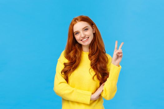 Waist-up shot cheerful tender and cute redhead modern woman in yellow sweater, tilt head and smiling lovely, showing peace sign, express positive emotions, joy and happiness, blue background.