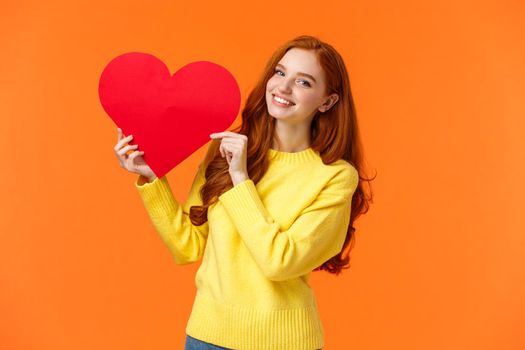 Valentines day perfect time to confess. Cute romantic and tender redhead woman in yellow sweater holding large heart sign and smiling, express affection and romance, standing orange background.