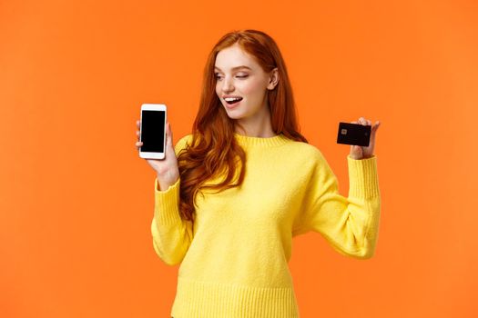 Enthusiastic pretty modern redhead girl advertising mobile banking, shopping online, holding smartphone and credit card, looking curious telephone screen, standing orange background.