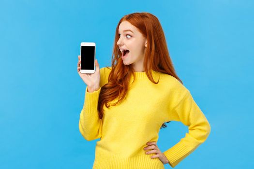 Wow look at this app. Amused and wondered, excited redhead girl looking at smartphone display, showing mobile screen, shopping site or application, standing blue background. Advertisement concept
