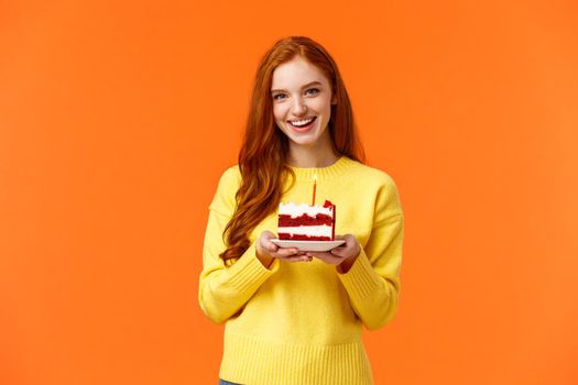 Girlfriend made surprise party, bring piece of cake with one candle to celebrate partner birthday, singing happy b-day and smiling lovely, standing orange background in yellow sweater.