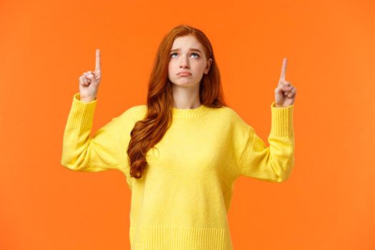 Sad and unhappy sulking redhead girl in yellow sweater, unsatisfied, look gloomy on christmas holidays, pointing and looking up envy or distressed, cant have desired gift, standing orange background.