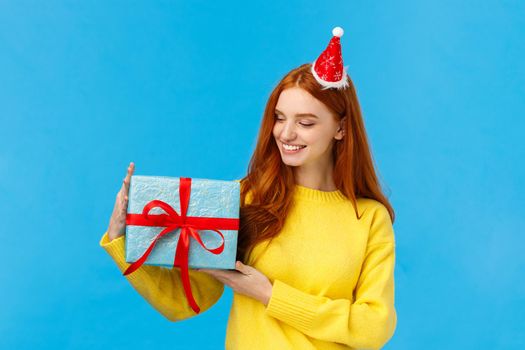 Holidays, love and family concept. Carefree tender and pretty redhead female holding cute wrapped box with gift, looking at present delighted, smiling happy, standing blue background joyful.