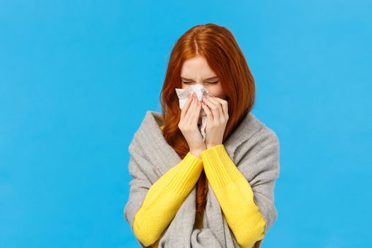 Seasonal flu. Cute redhead female student cought cold, sneezing in napkin, wear sweater and scarf on shoulders, feeling unwell sick, suffering high temprature, standing blue background.