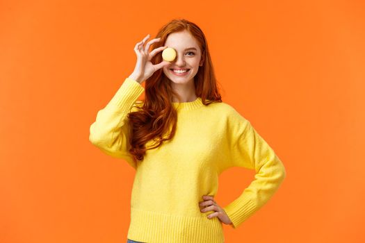 Forget about diet, lets enjoy. Cheerful and happy, smiling redhead woman holding small tasty macaron over eye and grinning, baking own desserts, promote awesome cafe with delicious food.