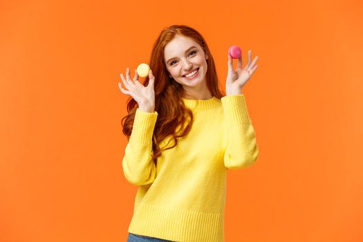 Girlfriend want share dessert, holding two macarons and smiling, suggest bite with lovely grin, tilt head flirty, standing happy orange background, enjoy winter holidays and sweet gifts.