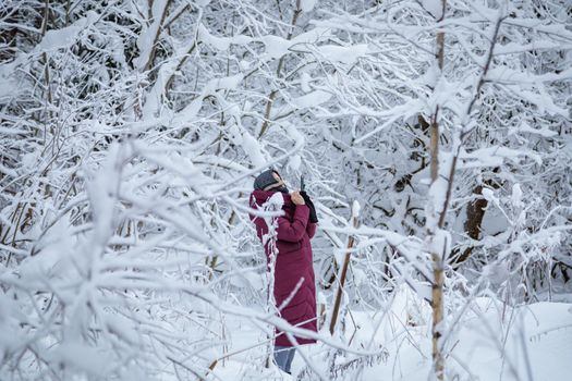 A girl in a burgundy down jacket photographs a snow-covered forest on a mobile phone.