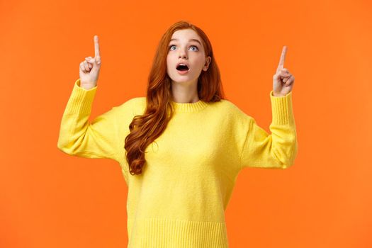 Excited, speechless and startled amazed redhead woman see awesome shopping sale promo, gaze and pointing up, stare top advertisement dropped jaw, breathtaking scenery, orange background.