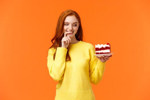 Cheerful and satisfied, happy redhead woman have cheat-day eating delicious food, holding tasty piece cake, biting lip and smiling, cant resist temptation, desire take bite, orange background.