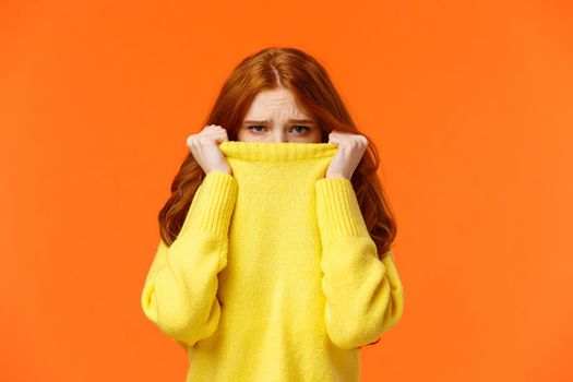 Cute timid tender redhead girl hate cold weather. Teenager sulking and frowning, looking unsatisfied, disappointed or lonely, pulling sweater on nose and staring camera offended, orange background.