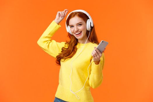 Lets dance. Cheerful carefree modern hipster girl with red curly hair and freckles, dancing lift hand up and holding smartphone, listen music with new ordered online headphones, orange background.
