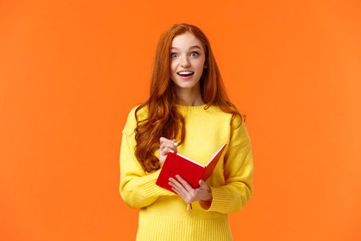 Inspired and upbeat, dreamy fascinated redhead girl writing down interesting lecture, smiling and gazing with admiration or enthusiasm, fill-in notebook schedule, standing orange background.