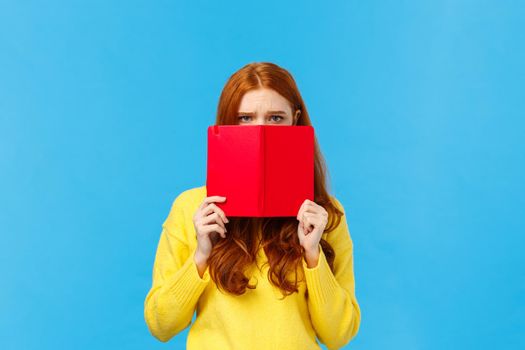 Upset gloomy and uneasy cute pouting redhead girl, frowning dont want speak to you, hiding face behind red notebook, frowning and staring offended, sulking over blue background, bad mood.