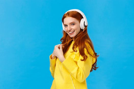 Coquettish and lovely cheerful pretty redhead woman with blue eyes, freckles, wearing white large headphones, posing profile and turn camera, rolling curl flirty smiling, standing blue background.
