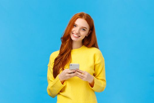Technology, internet and gadgets concept. Cute redhead woman sending text friend, messaging, having conversation using smartphone app, smiling camera, download useful application, blue background.