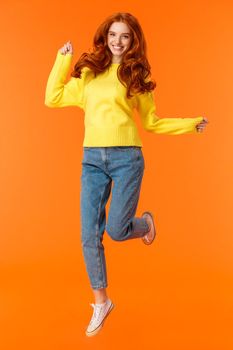 Vertical full-length portrait cheerful redhead excited girl jumping and feeling happy, smiling gazing camera carefree, cant wait open presents on winter holidays party, orange background.