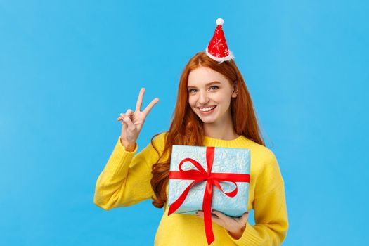Waist-up portrait of cute happy redhead woman receive gift in wrapped box, celebrating christmas holidays, wearing fancy new year hat and showing peace sign, smiling joyfully, blue background.