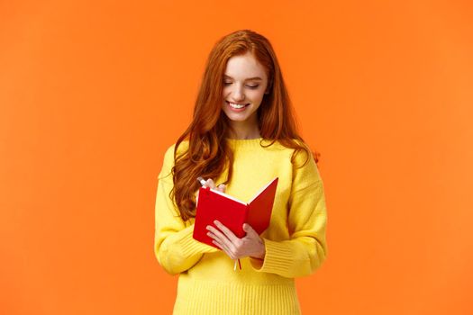 Waist-up portrait attractive redhead girl 20s write in her diary, fill-in to-do list or personal schedule in new notebook, smiling as having everything planned, standing orange background carefree.