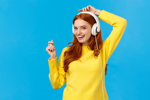 Cheeky and joyful, happy smiling redhead ecstatic woman having fun, listen music and lifting hands up as dancing, enjoying awesome sound quality, standing blue background delighted.