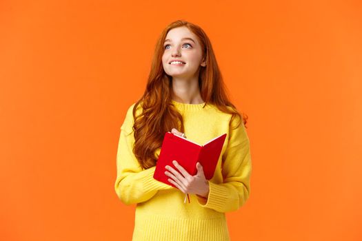 Dreamy, inspirational cute redhead female student writing daily tasks, to-do list in red lovely planner, look upper left corner thoughtful as smiling, getting inspiration to write novel, orange wall.