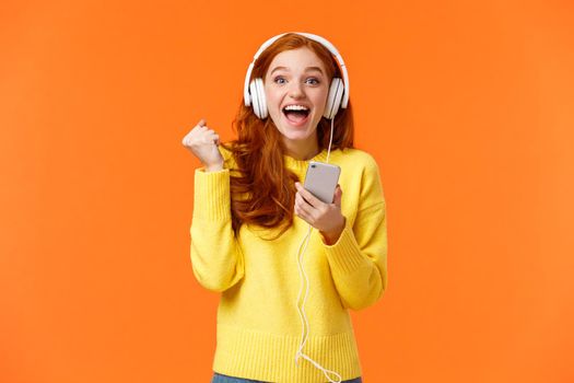 Hooray yes new song. Attractive cheerful and excited redhead woman fist pump in joy and positive emotions, wearing headphones, holding smartphone, smiling camera happily, orange background.