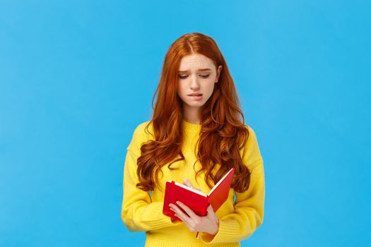 Perplexed and uneasy, concerned cute redhead female student having troubles with schedule, frowning reading girlfriend diary, feeling guilty, holding red notebook, feeling troubled, blue background.