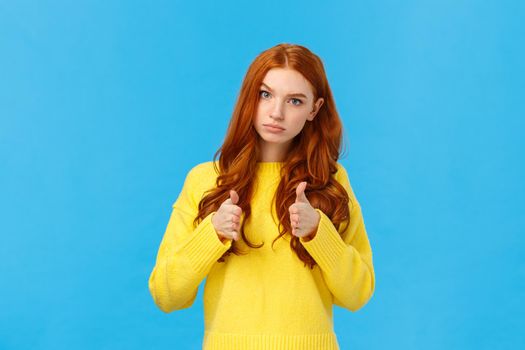 Serious and determined supportive redhead girlfriend, showing you can do it, encourage friend, make thumbs-up in approval, rooting for you, glance with confident expression, blue background.