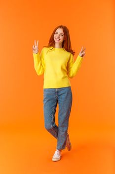 Vertical full-length cute lovely redhead teenage girl in jeans and winter yellow sweater standing with peace signs over orange background, smiling posing express carefree joy emotions.