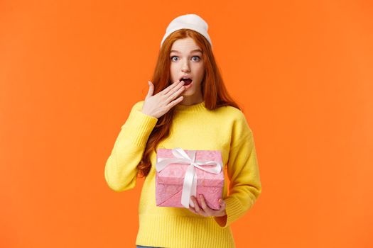 Girl surprised didnt expact gift from you. Amused and startled cute redhead girlfriend open mouth, cover it with hand as receive cute pink box of present, look camera excited, orange background.