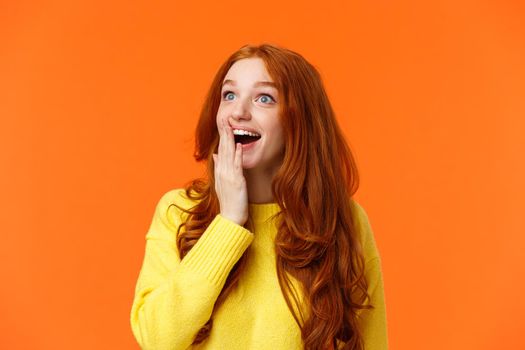 Fascinated and amiring cute redhead woman looking impressed and excited at upper left corner, smiling see breathtaking view, cover opened mouth palm thrilled, losing speech happy, orange background.