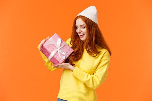 Girl trying figure-out whats inside gift box shaking it and looking intrigued. Excited happy redhead woman like winter holidays and receiving present, standing amused orange background.