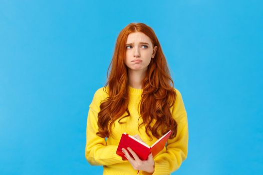 Troubled and pensive uneasy pretty redhead female, pouting and frowning looking away thoughtful, feeling sad and distressed, reading something bad in notebook, standing blue background.