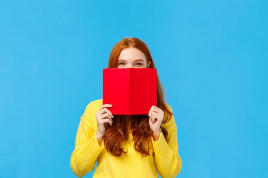 Sassy and creative happy charismatic redhead woman, teenager hiding face behind red notebook and smiling, grinning sly, hide from friend what she wrote in diary, standing blue background.
