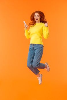 Vertical full-length shot carefree, excited and pleased good-looking ginger girl in sweater, jeans, jumping over orange background, holding smartphone show thumbs-up and smiling approvingly.