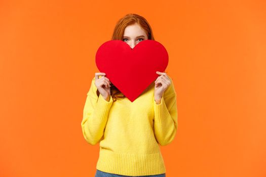 Cute romantic and blushing girl express her feelings on valentines day, hiding face shy behind big heart sign and peeking at camera, giving lovely gift, standing orange background tender.