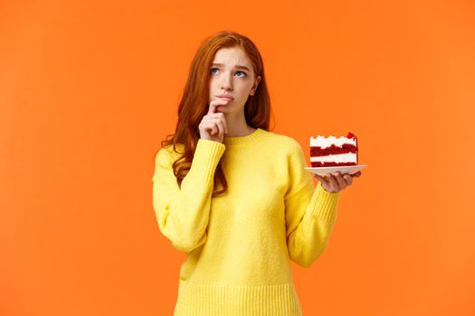 Worried, concerned silly young redhead girl taking care her body, calculating calories, being diet and want eat delicious cake, holding piece of dessert, look up thoughtful and troubled, orange wall.